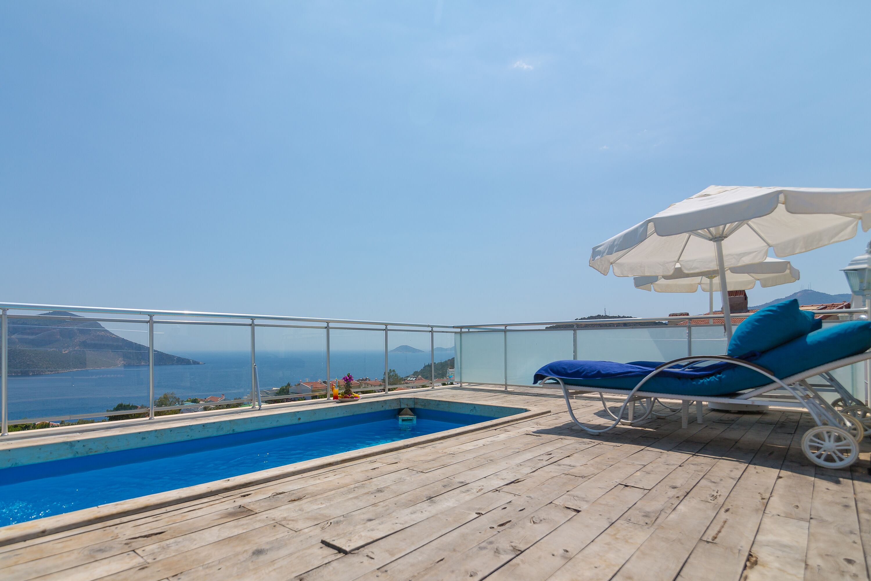 Una terraza de madera con piscina, una silla de descanso y una sombrilla blanca.