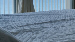 Un lit recouvert d’une couverture blanche à motifs, une vue sur la mer par la fenêtre et une rambarde blanche en arrière-plan.