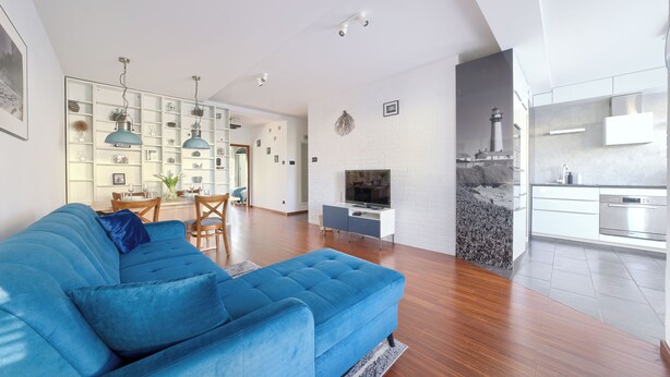 Ein modernes Wohnzimmer mit einem blauen Sofa, einem Essbereich mit einem Holztisch und einer Küche mit weißen Schränken und einem Edelstahlbackofen.