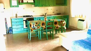 A kitchen with green cabinets, a dining table with sunflower-patterned chairs, and a bed with a blue patterned bedspread. - Fantastico San Teodoro STRAU02 (San Teodoro)