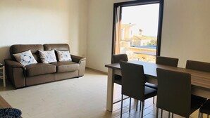 A living room with a brown leather sofa, two patterned cushions, a dining table with four chairs, and a large window offering a view of buildings and a balcony.