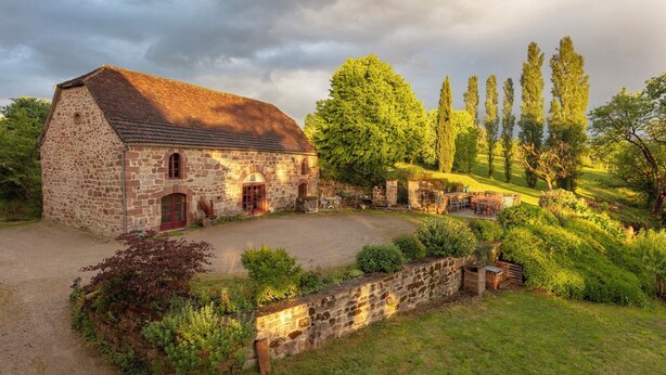 Ein Steinhaus mit roter Tür, einem gepflasterten Auffahrt und einem Garten mit Bäumen und Sträuchern.
