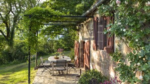 Eine Terrasse im Freien mit Steinböden, einem runden Tisch und Stühlen, umgeben von Grünpflanzen und einer Steinmauer.
