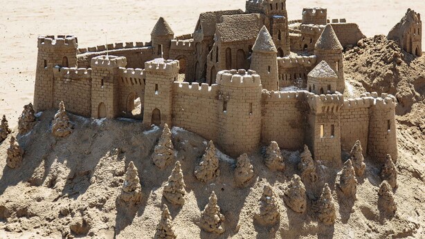 Un castillo de arena con múltiples torres y murallas, situado en una playa arenosa con gente y sombrillas al fondo.