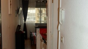 Un pasillo estrecho con una cocina visible a través de una puerta abierta. Hay un teléfono en la pared, un radiador y una ventana con cortinas.