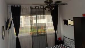 Un dormitorio con cama, ventilador de techo, ventana con cortinas, espejo y cómoda.