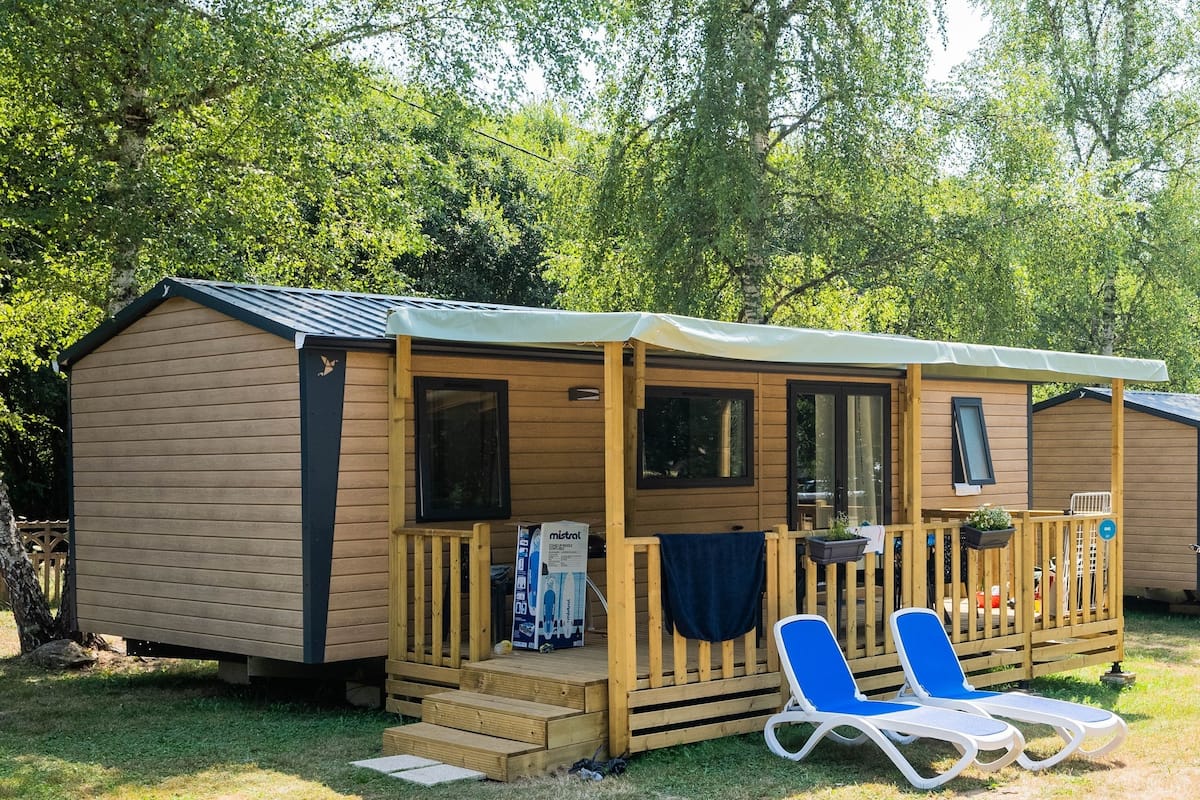 A wooden mobile home with a porch, two blue lounge chairs, and a small garden area.
