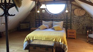 Quarto de sótão com cama, vigas de madeira, parede de pedra, claraboia circular e ventoinha de teto.