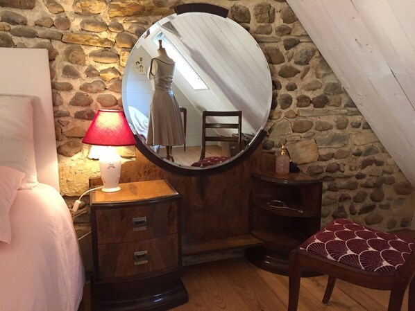 A bedroom with a stone wall, a round mirror, a wooden dresser, a bedside table with a lamp, a chair, and a bed. - B&B Maison D'Orride (Ledeuix)
