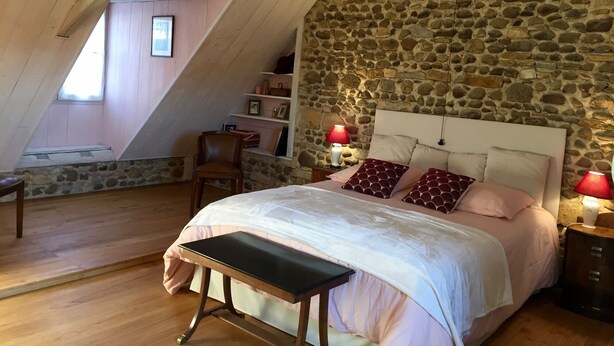 A bedroom with a stone wall, a bed with pink bedding, a wooden bench, and a nightstand with a lamp. - B&B Maison D'Orride (Ledeuix)