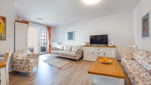A living room with a sofa, a coffee table with fruit, a TV, and a view to the outside. - Apartamenty Sun & Snow Na Solnej (Kolobrzeg)