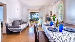 Ein modernes Wohnzimmer mit einer Couch, einem Esstisch und Stühlen. Auf dem Tisch steht ein Fernseher und eine Pflanze. Das Zimmer bietet einen Blick ins Freie.