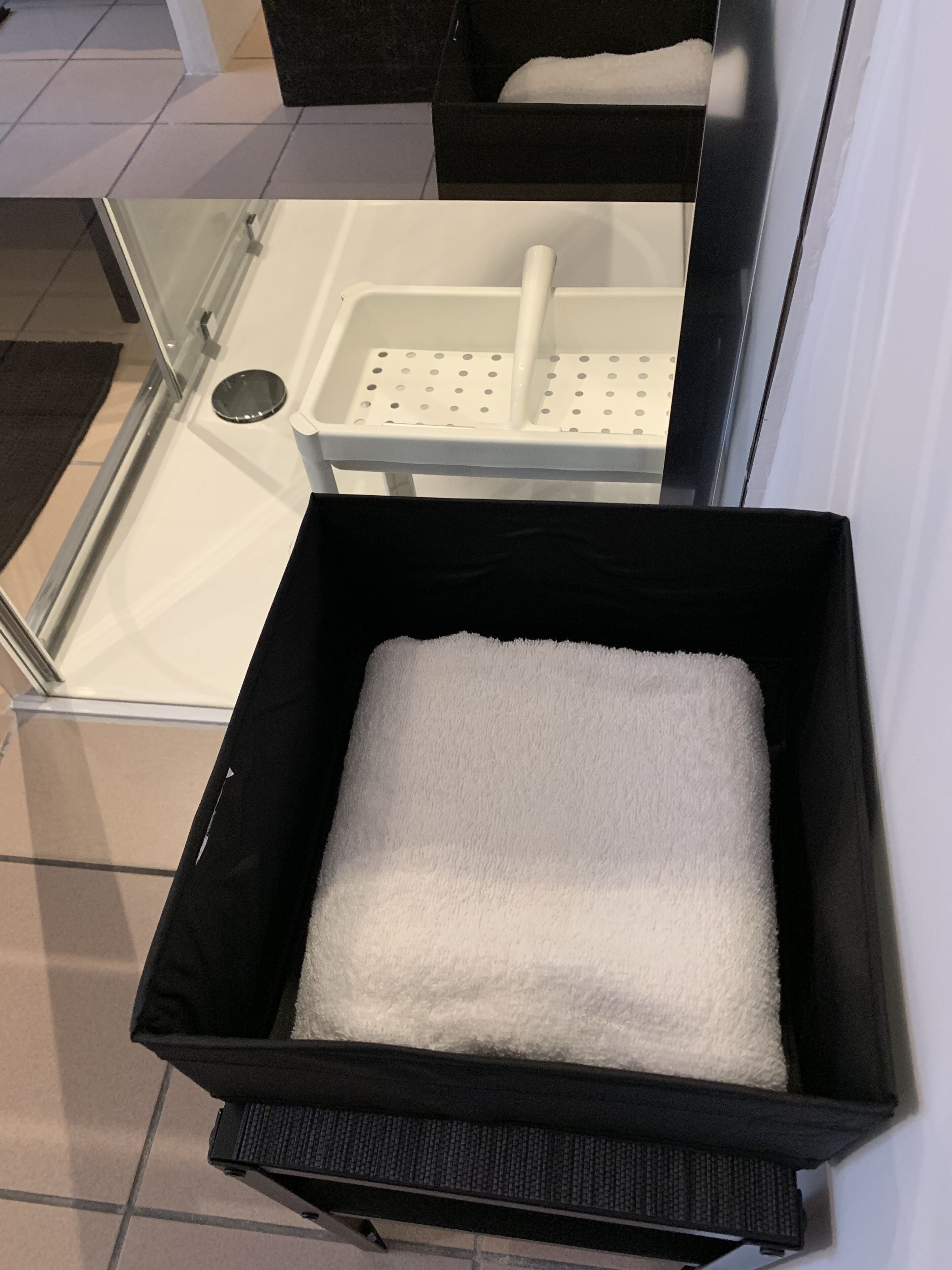 Una caja de almacenamiento negra con una toalla blanca en su interior, sobre un piso de baldosas cerca de una zona de ducha con mampara de vidrio.