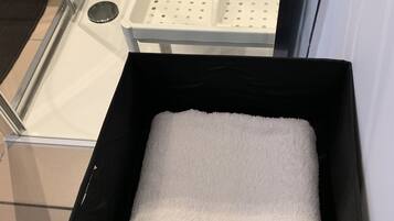 Una caja de almacenamiento negra con una toalla blanca en su interior, sobre un piso de baldosas cerca de una zona de ducha con mampara de vidrio.