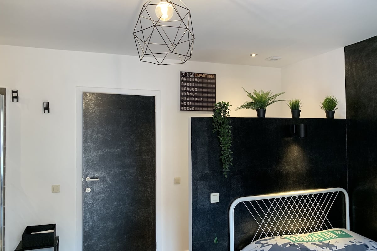 A modern bedroom with a geometric light fixture, a black door, and a sign listing departure destinations.