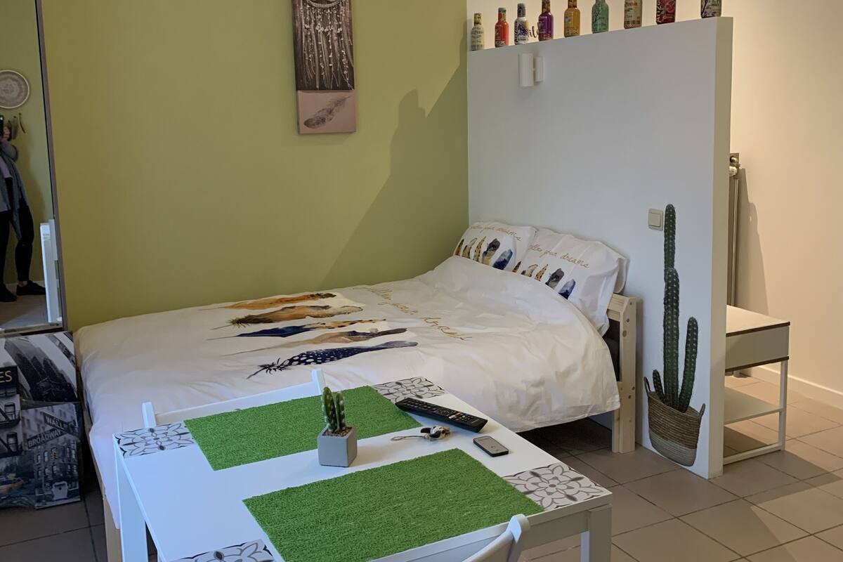 A bedroom with a bed, a table with a cactus, a chair, a wall-mounted shelf with bottles, and a hanging light fixture.