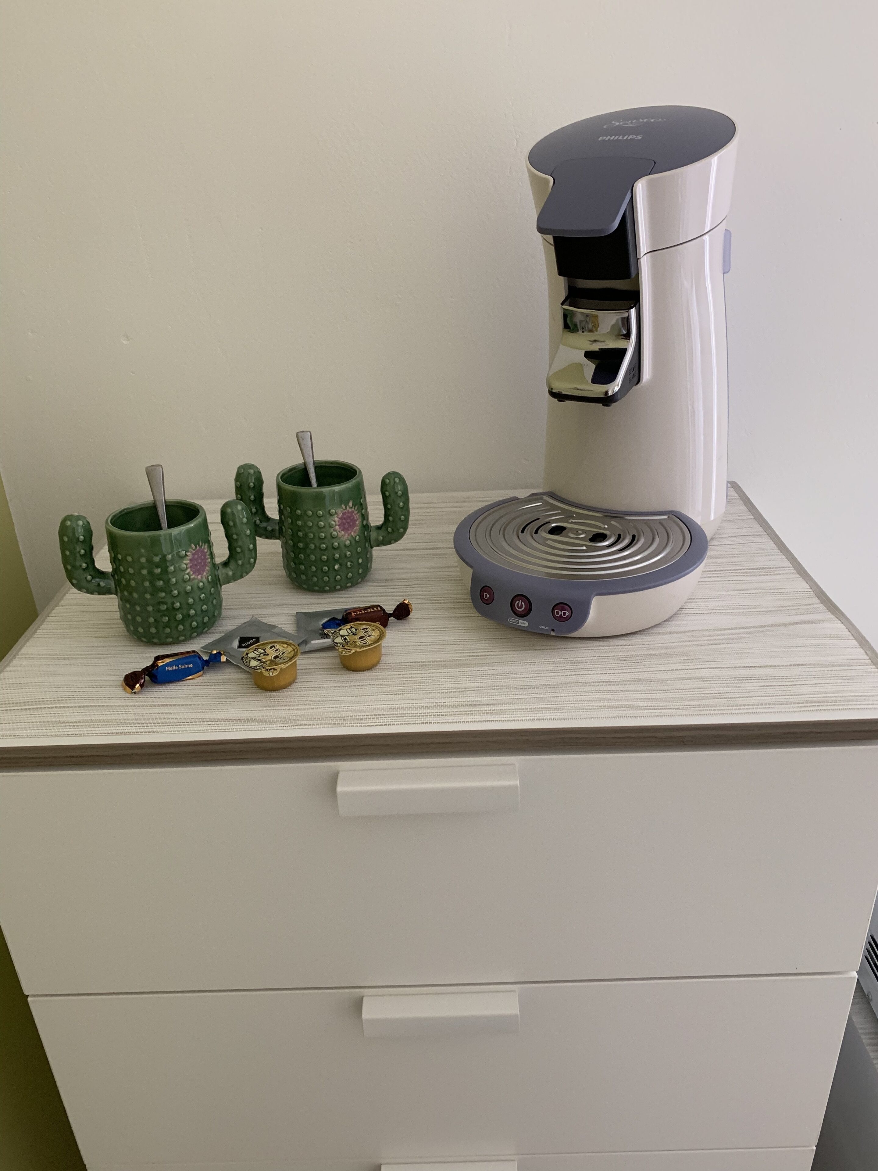 A white drawer with a coffee machine, two cactus-shaped mugs, and some chocolates on top.