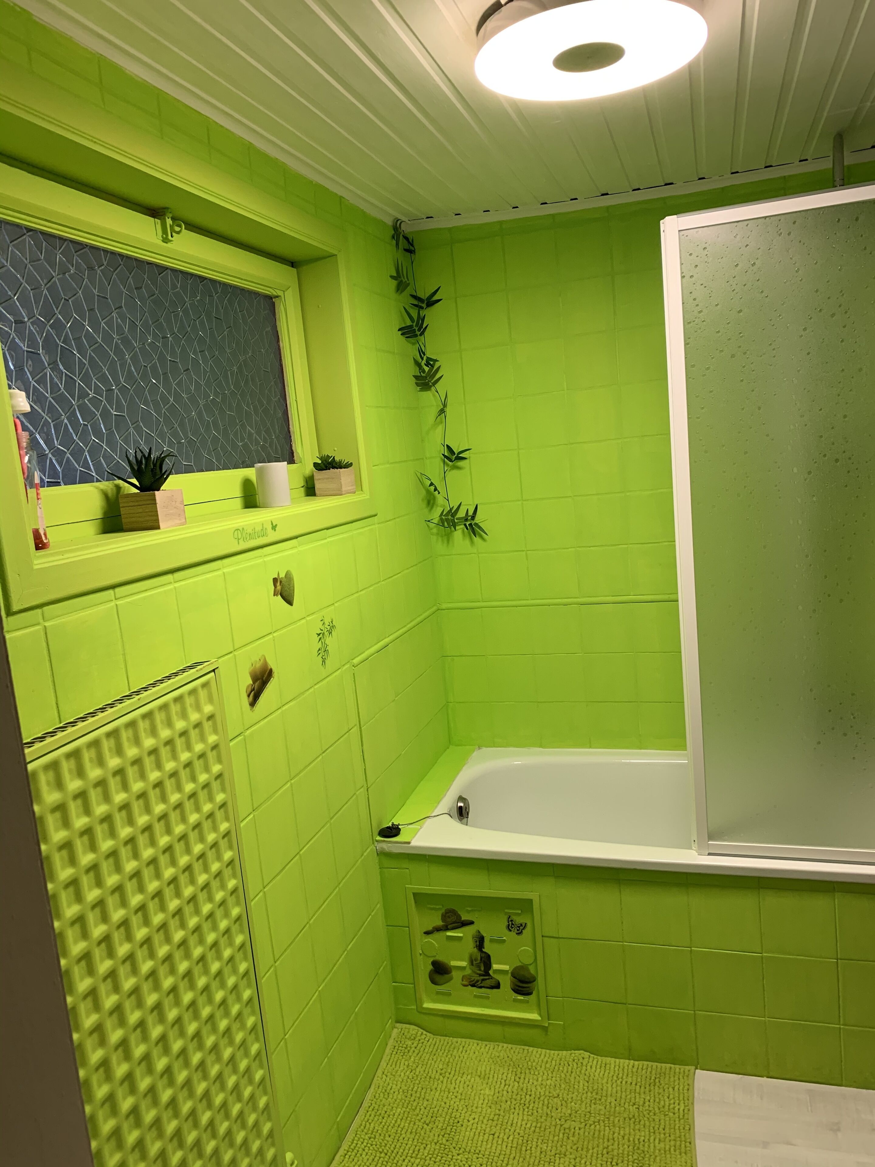 Een badkamer met groene tegels, een raam met planten, een bad en een toilet.