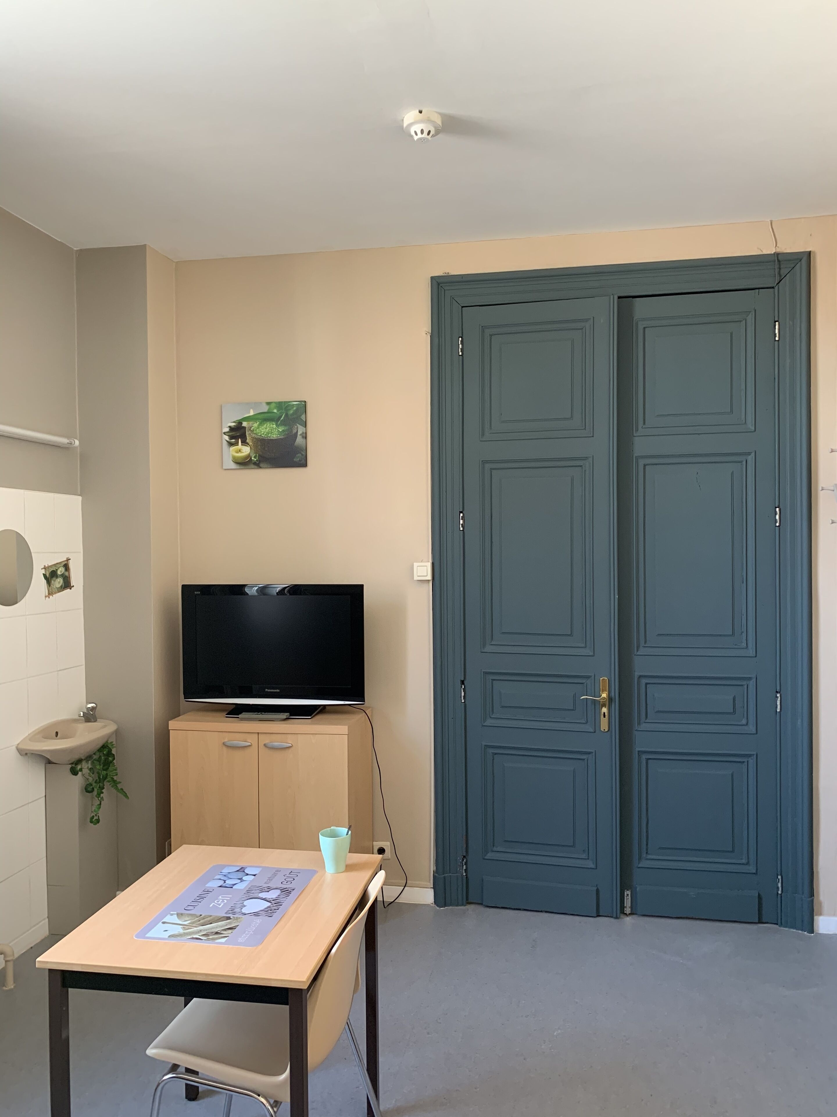 Ein Zimmer mit Fernseher auf einer Holzkommode, einem Tisch mit Platzdeckchen, einem Waschbecken und zwei Türen.