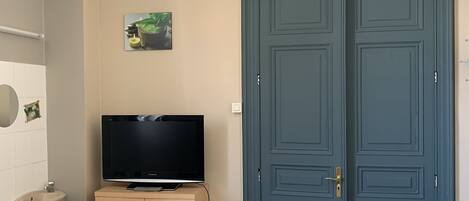 A room with a TV on a wooden cabinet, a table with a placemat, a sink, and a pair of doors.