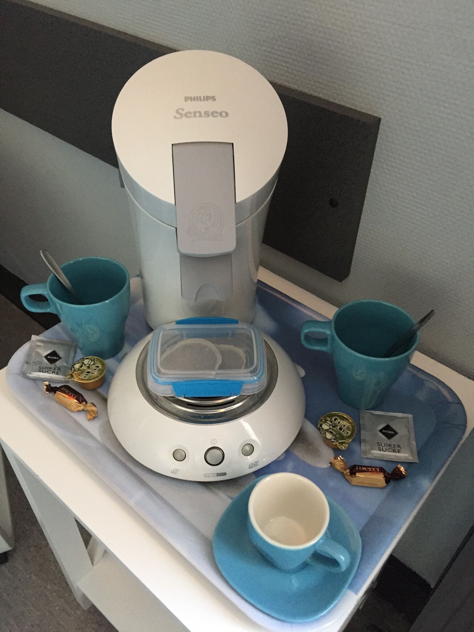 Una cafetera con una taza de café, dos tazas adicionales y algunos aperitivos sobre una mesa.