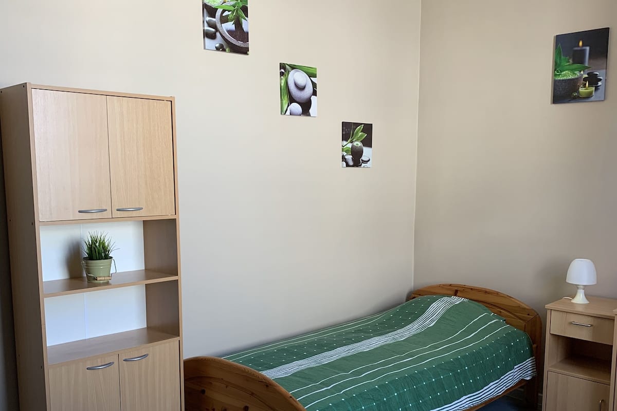 A small, neatly arranged bedroom with a single bed, a wooden desk, a chair, a small cabinet, and a nightstand.