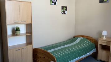 A small, neatly arranged bedroom with a single bed, a wooden desk, a chair, a small cabinet, and a nightstand.