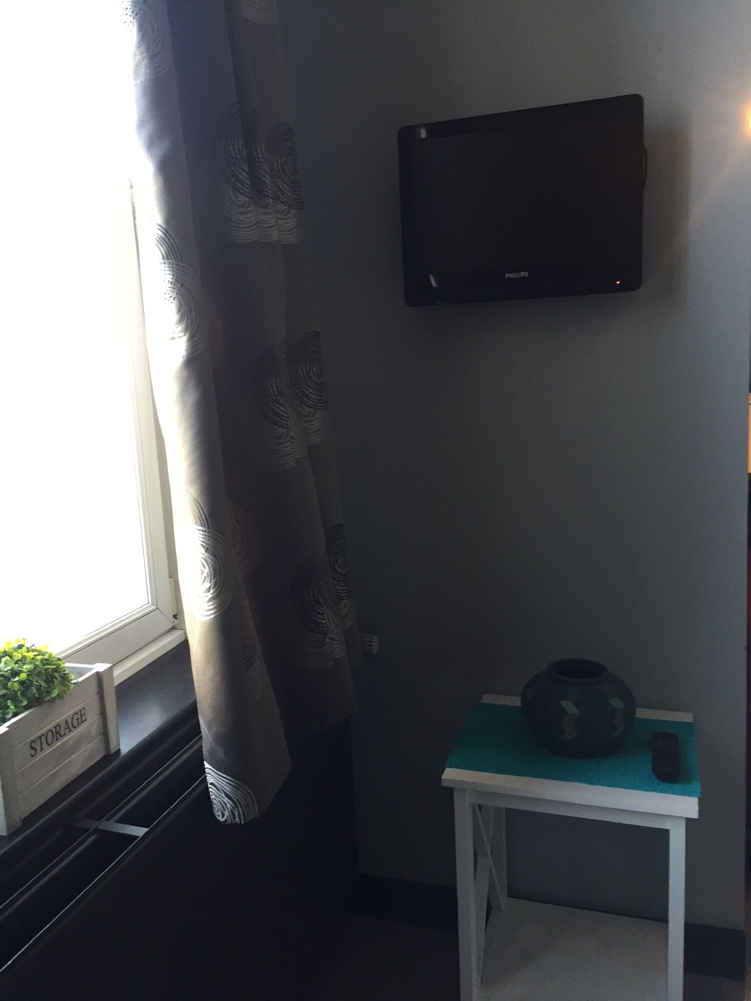 Una habitación con ventana, televisor montado en la pared, una pequeña mesa con un adorno y un mueble de almacenamiento etiquetado 'STORAGE' en el alféizar.