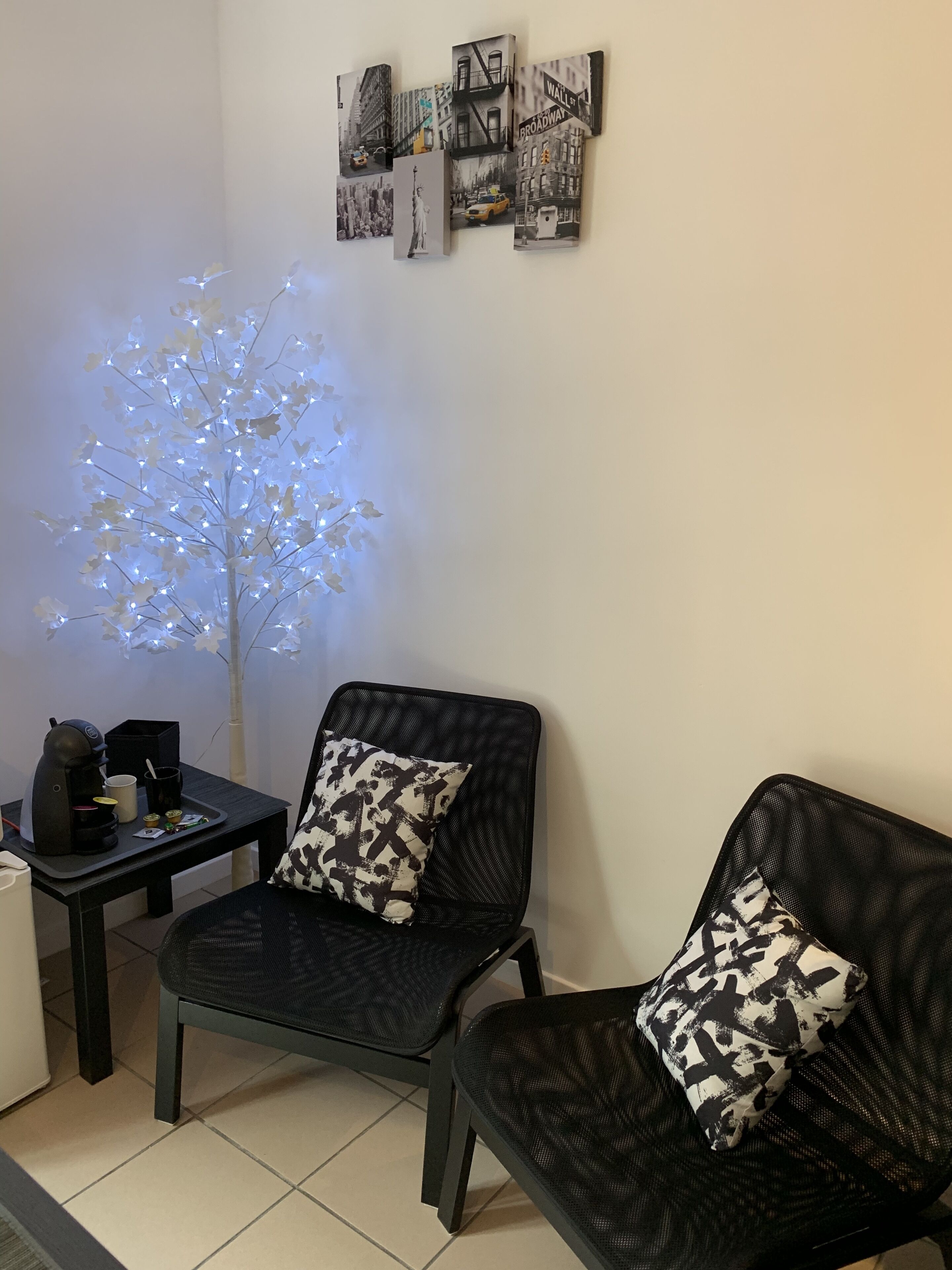 A modern living room with a coffee machine, two black chairs with patterned cushions, a small table, and a wall-mounted light tree.