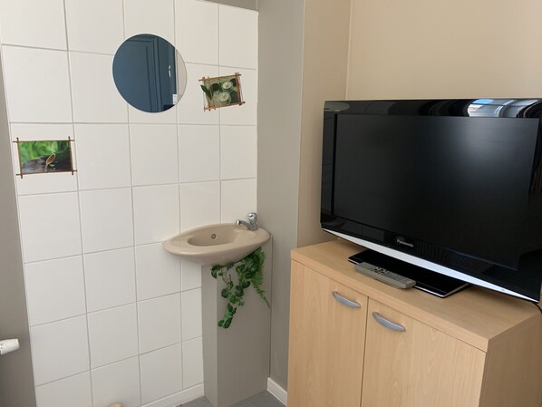 Un baño con inodoro, lavamanos, espejo y televisor sobre un mueble de madera.