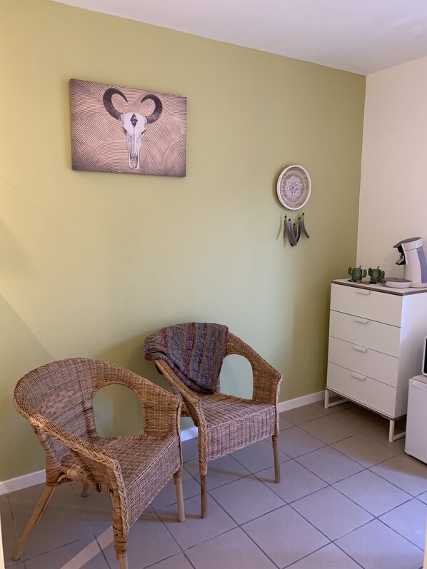 Una habitación con dos sillas de mimbre, un tocador blanco, una cafetera y una obra de arte en la pared.