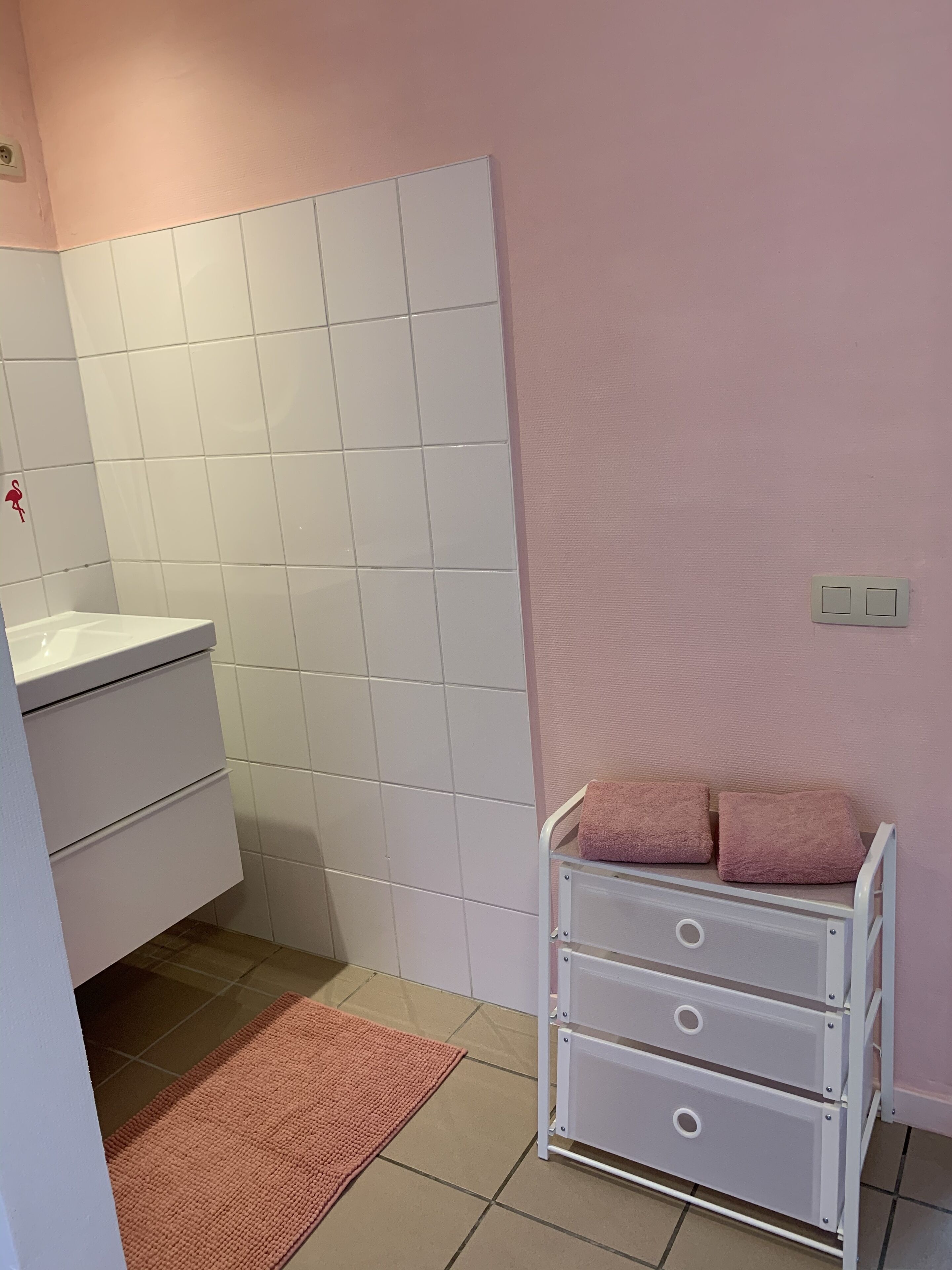 A bathroom with white tiles, a pink wall, a white sink, a white cabinet, and a white towel rack with towels.