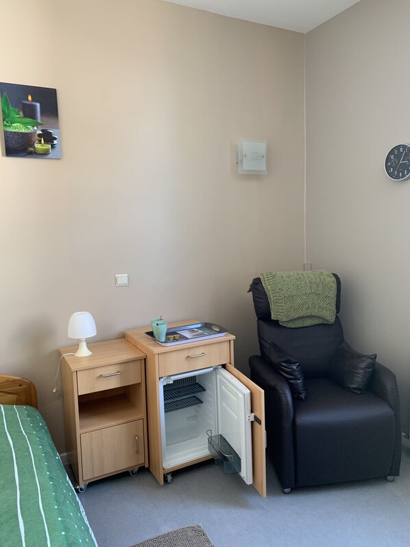 A compact room with a bed, a desk with a lamp, a chair with a blanket, a wall clock, and a small refrigerator.