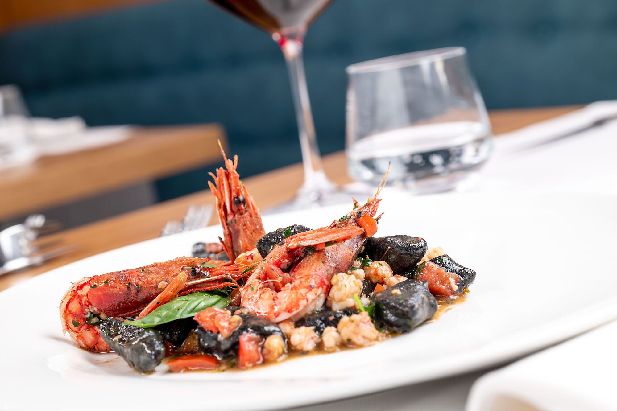 A seafood dish on a white plate with a glass of water and a glass of red wine on the table.