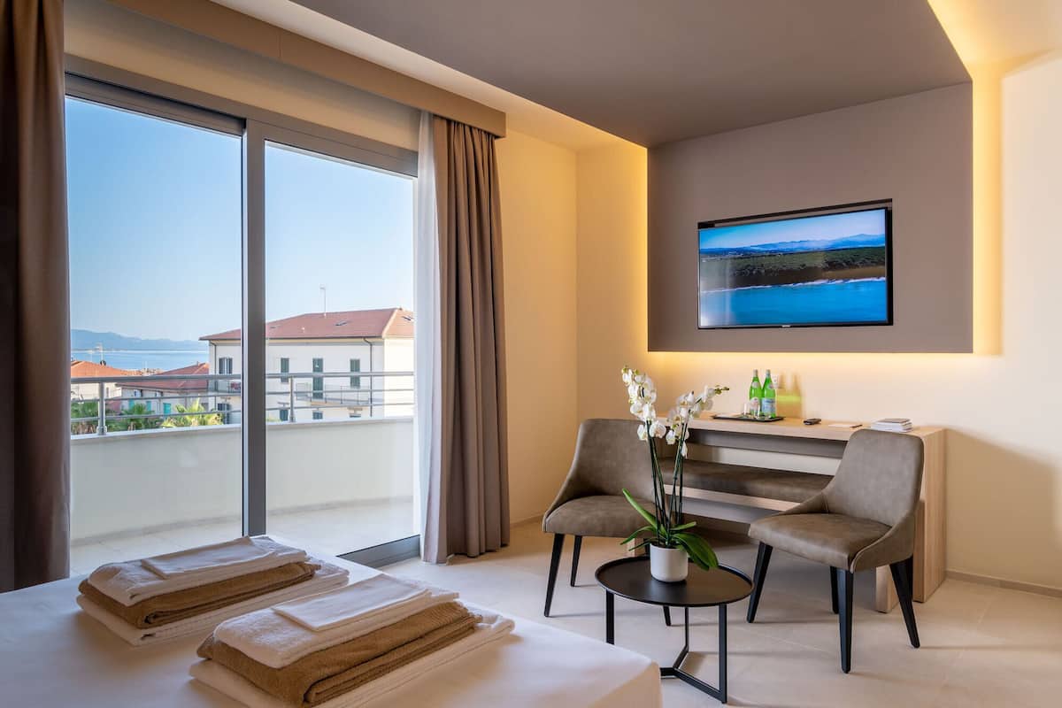 A hotel room with a large window, a bed with towels, two chairs, a small table, a TV, and a view of the sea and buildings.