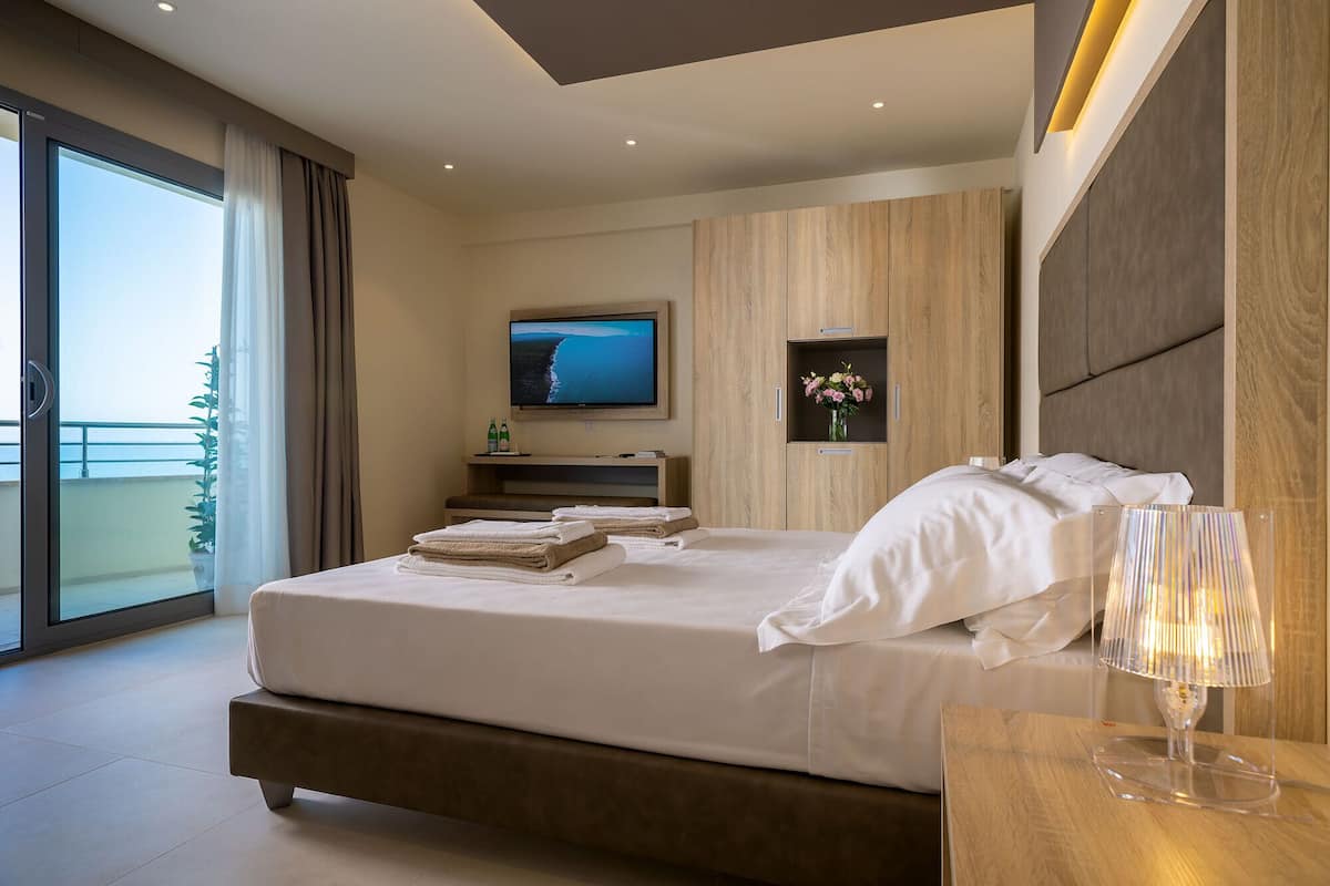 A modern hotel room with a large bed, a TV, a bedside table with a lamp, and a view of the sea through the balcony.
