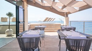 Un área de comedor al aire libre con mesas y sillas, una planta grande en maceta y vistas al mar.