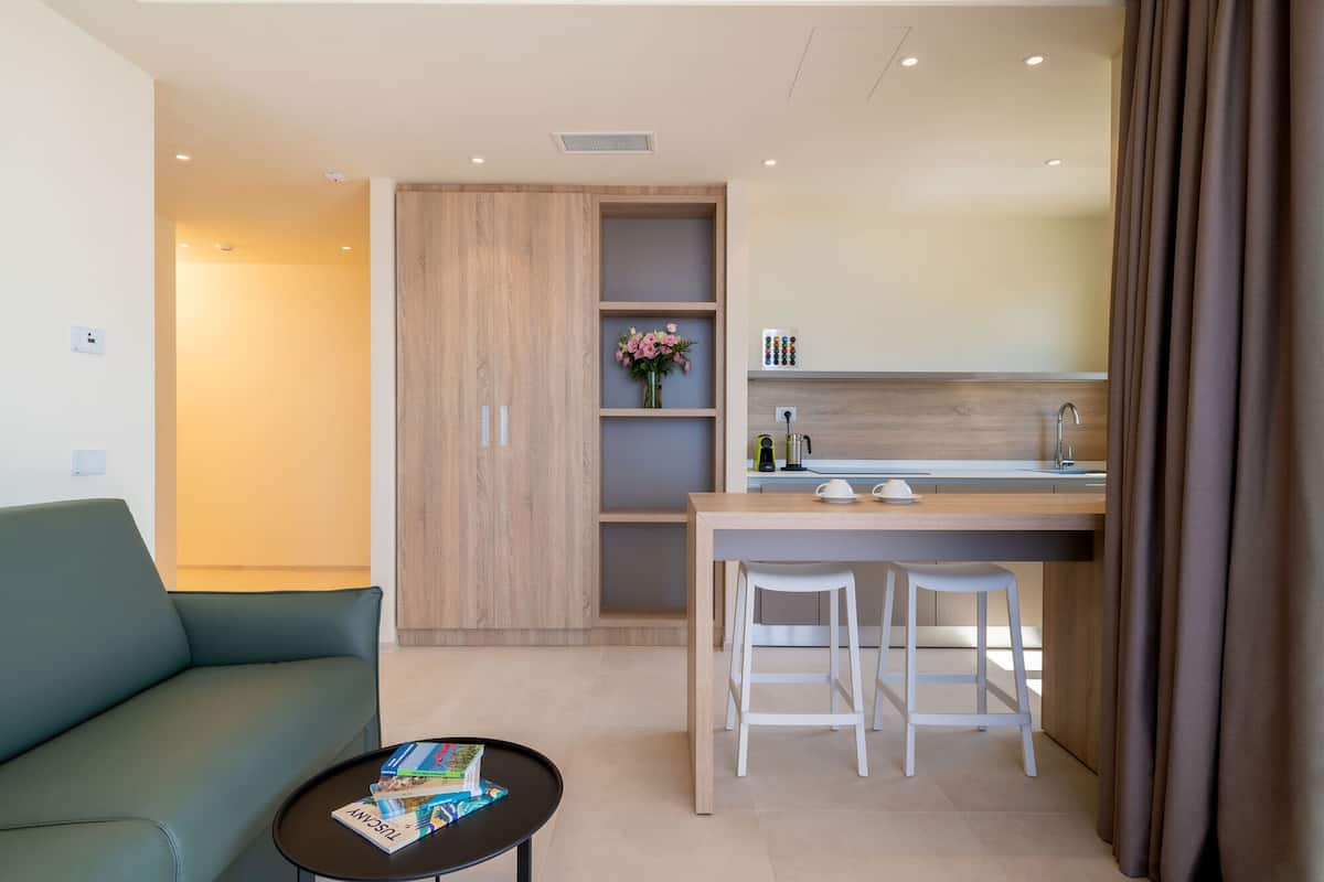A compact living space with a sofa, a small coffee table, a kitchenette with a sink and bar stools, and a wooden wardrobe.