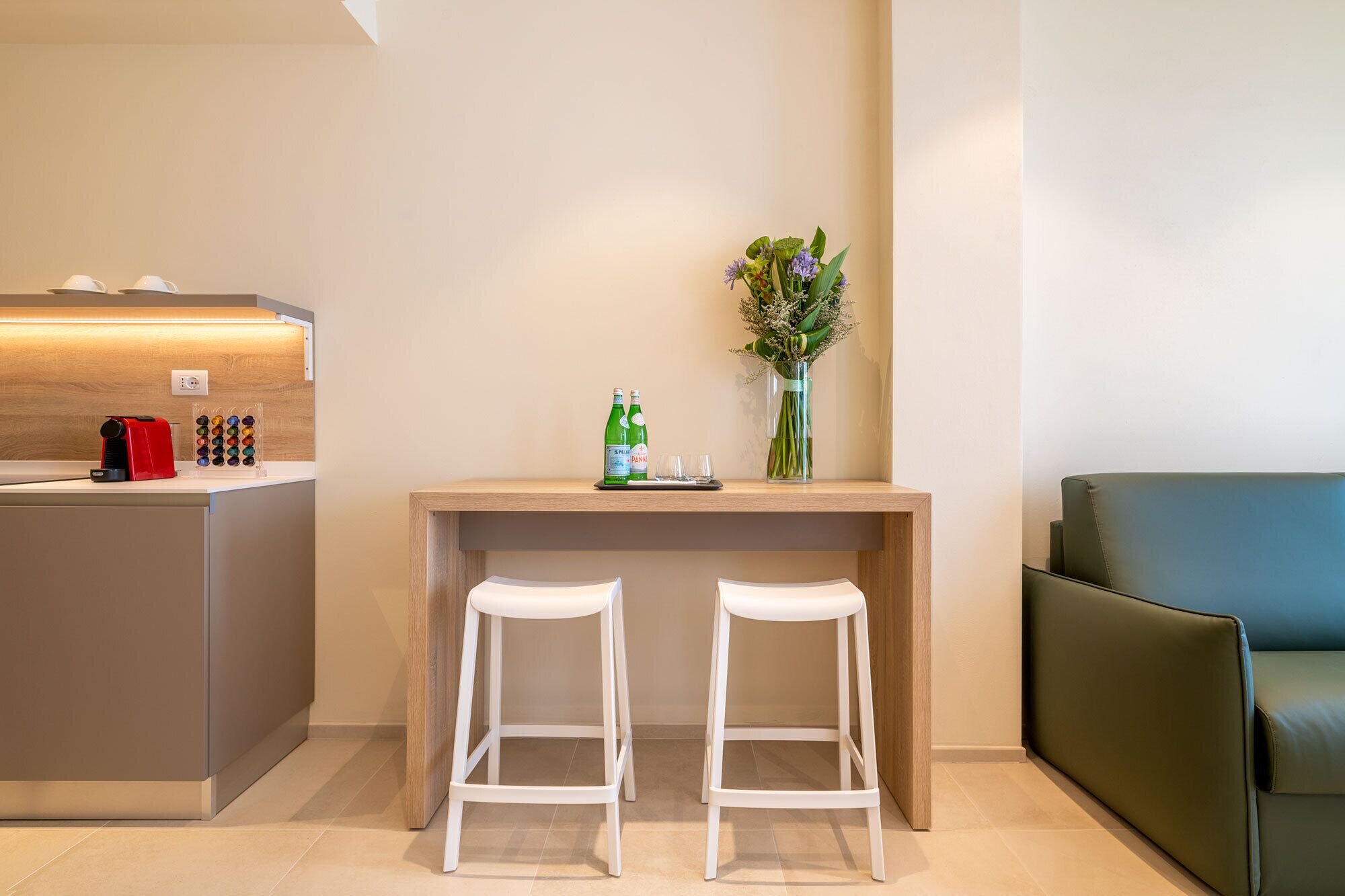 A modern kitchen with a coffee machine, two stools, a small table with drinks, and a vase with flowers.