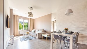 A modern living room with a sofa, dining table, and chairs. There is a TV mounted on the wall and a window with curtains. - Apartamenty Sun&Snow Polanki Park (Kolobrzeg)