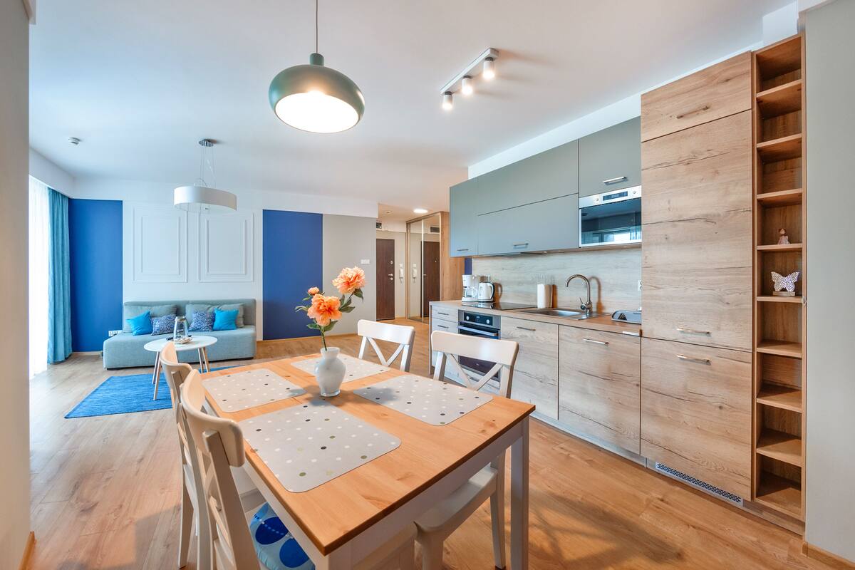 A modern kitchen with wooden cabinets, a dining table with chairs, and a blue sofa in the living area.