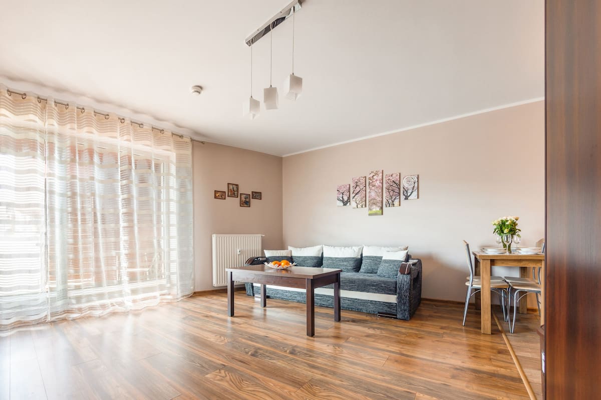 A living room with a wooden floor, a sofa, a coffee table, a dining table with chairs, and a radiator.