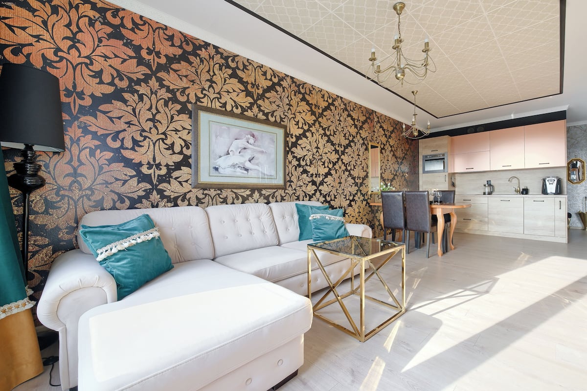 A modern living room with a white sofa, teal cushions, a glass coffee table, and a patterned wallpaper. A dining area with a wooden table and chairs is visible in the background.