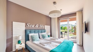 A modern bedroom with a large bed, a headboard with 'Sea & Sky' sign, a bedside table with a lamp, and a view of the outdoors through a sliding glass door. - Apartamenty Sun&Snow Polanki Park (Kolobrzeg)