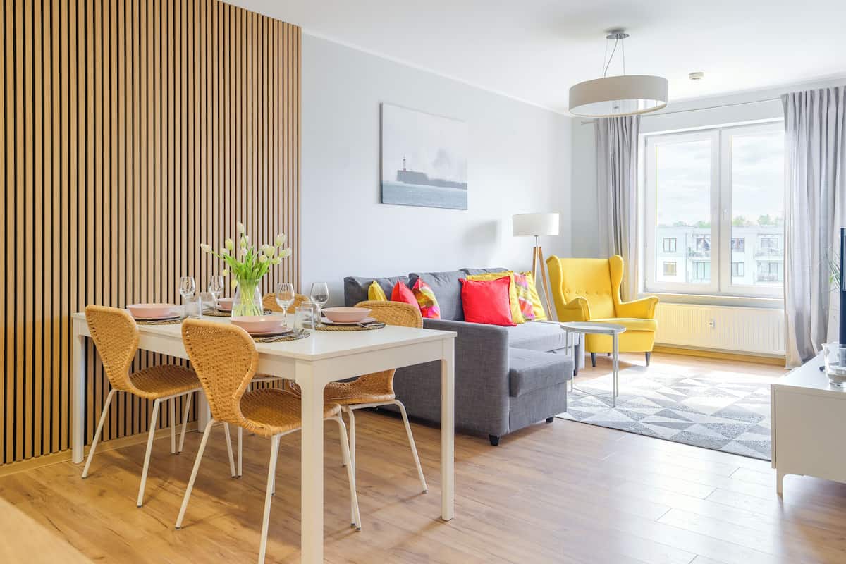 A modern living room with a dining area, featuring a wooden slat wall, a grey sofa with colorful cushions, a yellow armchair, and a large window with a view of buildings.