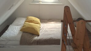 Une petite chambre mansardée de forme triangulaire, avec un lit simple, une lucarne et une échelle en bois.