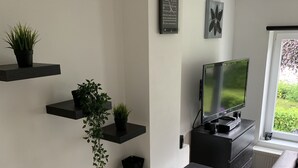A modern living room with a flat-screen TV, a built-in oven, and black floating shelves with plants. - Petit Coin De Paradis (Charleroi)