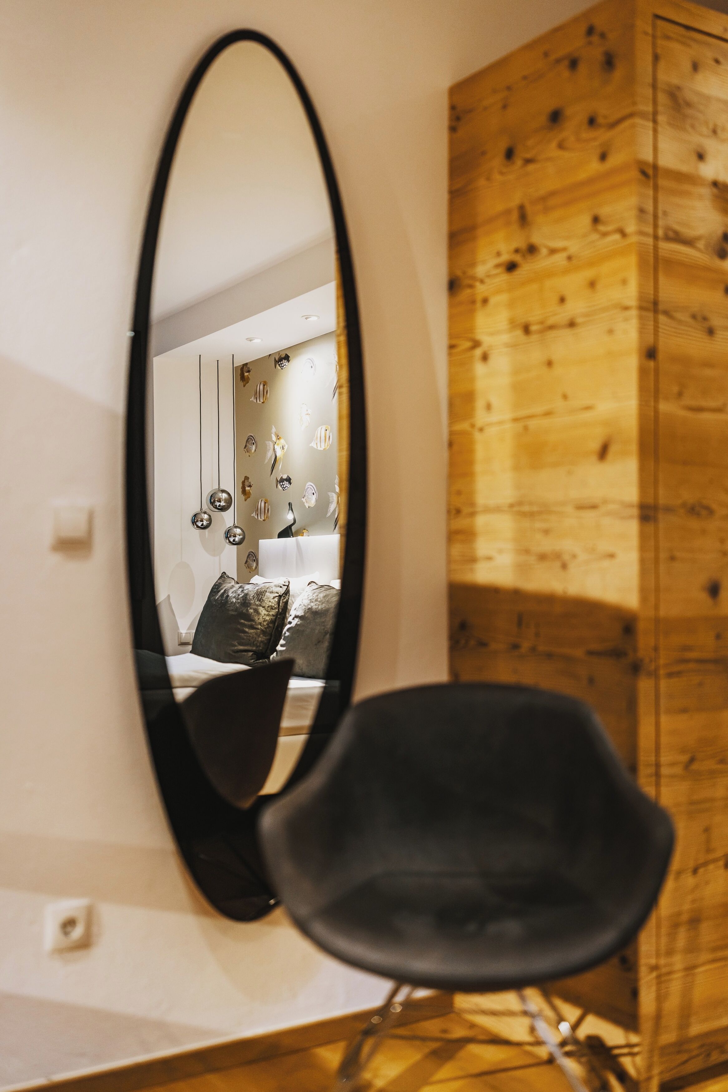 Une pièce comprenant une armoire en bois, une chaise noire et un grand miroir qui reflète un lit doté d’une tête de lit blanche et agrémenté d’œuvres d’art décoratives sur le mur.