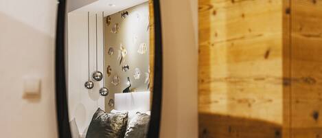 Une pièce comprenant une armoire en bois, une chaise noire et un grand miroir qui reflète un lit doté d’une tête de lit blanche et agrémenté d’œuvres d’art décoratives sur le mur.