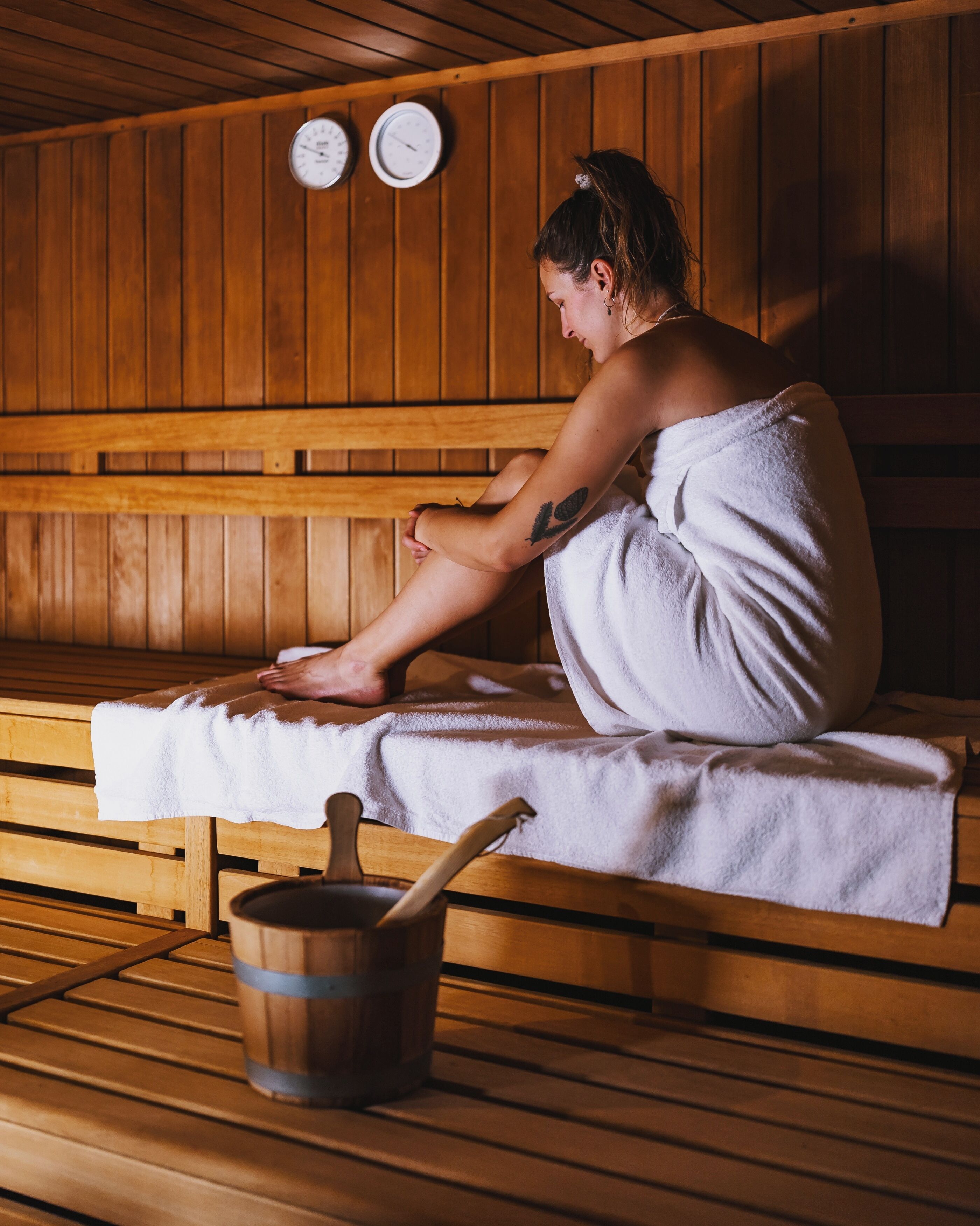 Una persona sentada en un banco dentro de un sauna de madera, con una toalla alrededor del cuerpo. Hay dos relojes en la pared y un balde de madera con una cuchara.